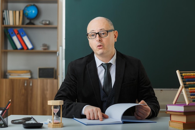 Man teacher wearing glasses checking class register looking puzzled sitting at school desk in front of blackboard in classroom