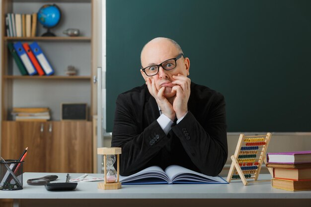 Man teacher wearing glasses checking class register looking tired and bored making grimace sitting at school desk in front of blackboard in classroom