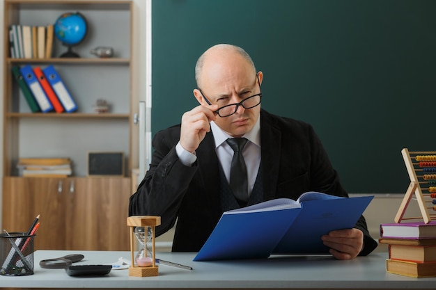 教室の黒板の前の学校の机に座って懐疑的な表情で見ているクラスレジスタをチェック眼鏡をかけている男の先生