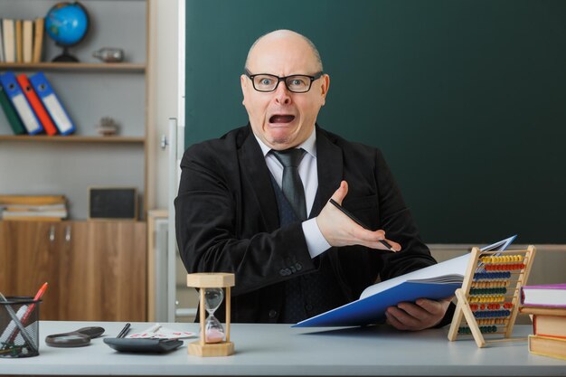 Man teacher wearing glasses sitting at school desk checking class register in front of blackboard in classroom looking at camera in panic making wry mouth