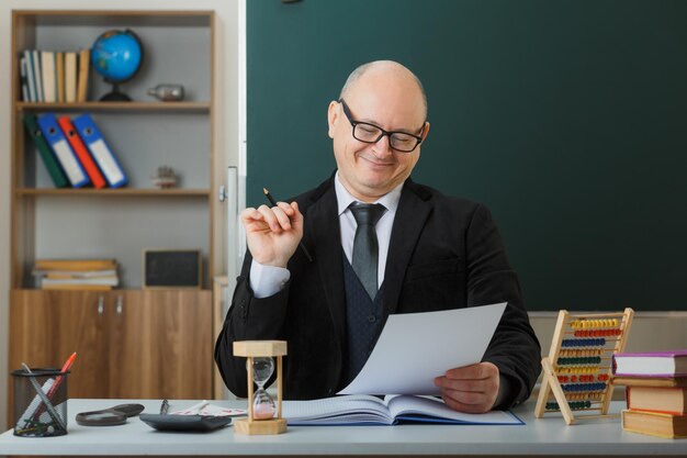 Man teacher wearing glasses sitting at school desk in front of blackboard in classroom checking homework of students happy and pleased smiling