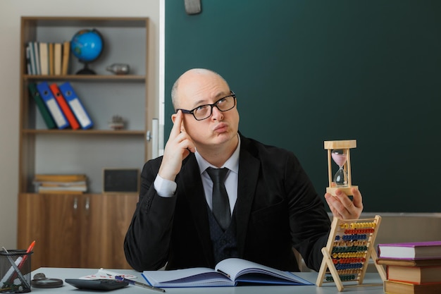 man teacher wearing glasses sitting at school desk in front of blackboard in classroom holding hour glass explaining lesson looking up puzzled