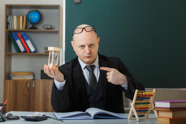 Man teacher wearing glasses sitting at school desk in front of blackboard in classroom holding hour glass explaining lesson pointing with index finger with serious face