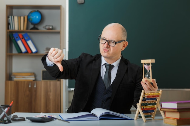 教室の黒板の前にある学校の机に座って眼鏡をかけている男の先生が時間のガラスを持って親指を下に向けてレッスンを説明している