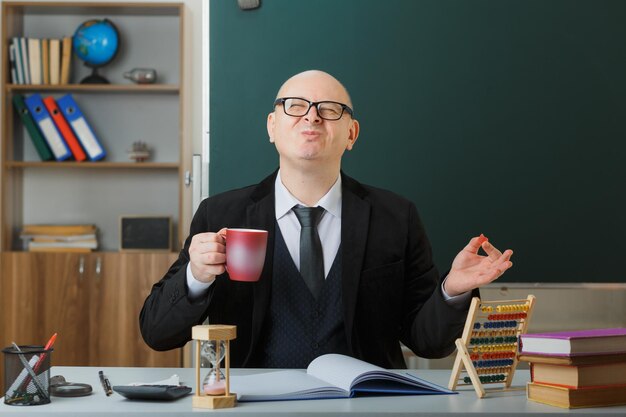 Man teacher wearing glasses sitting at school desk in front of blackboard in classroom holding mug of coffee happy and pleased smiling