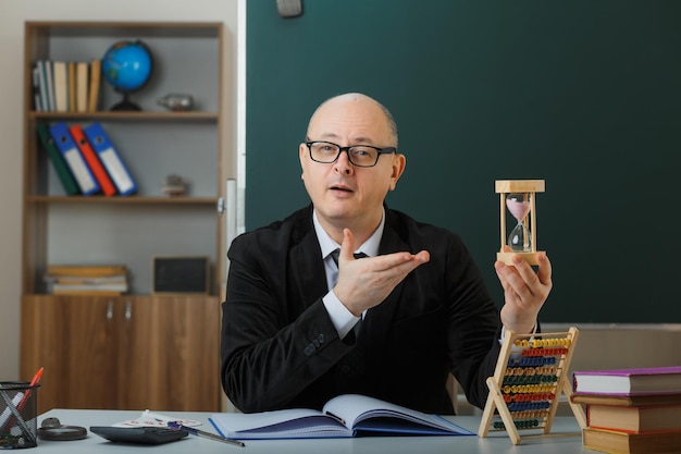 Man Teacher Wearing Glasses Sitting At School Desk In Front Of Blackboard In Classroom Presenting Hour Glass Explaining Lesson Looking Confident