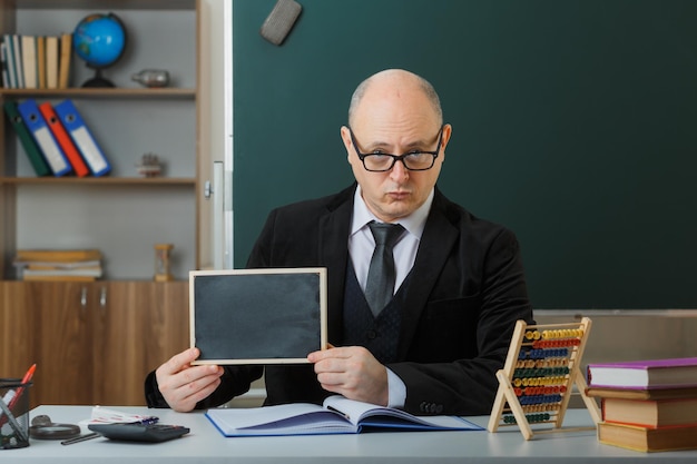 Man teacher wearing glasses sitting at school desk in front of blackboard in classroom showing chalkboard explaining lesson explaining lesson with serious face