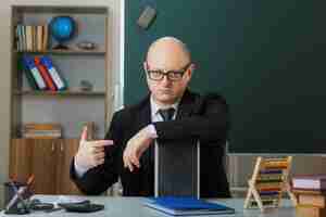 Free photo man teacher wearing glasses sitting at school desk in front of blackboard in classroom showing chalkboard explaining lesson looking displeased