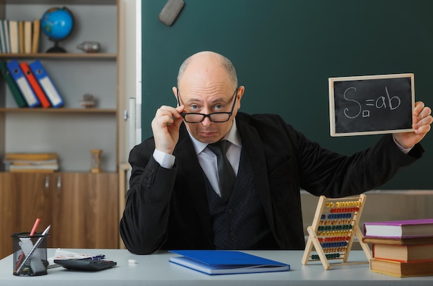 Foto gratuita insegnante uomo con gli occhiali seduto al banco di scuola davanti alla lavagna in classe che mostra la lavagna che spiega la lezione che sembra sorpreso