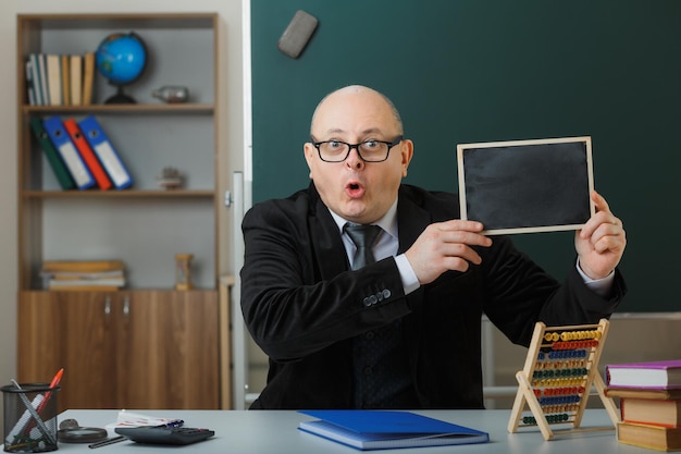 Man teacher wearing glasses sitting at school desk in front of blackboard in classroom showing chalkboard explaining lesson looking surprised