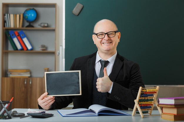 教室の黒板の前の学校の机に座って眼鏡をかけている男の先生は、笑顔で親指を示すレッスンを説明する黒板を示しています