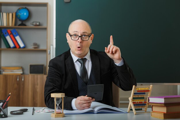 Учитель-мужчина в очках сидит за школьной партой с классным журналом перед доской в классе, объясняя урок, держа калькулятор, удивленно указывая указательным пальцем вверх