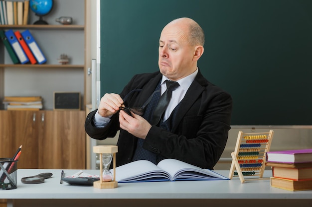 Free photo man teacher wearing glasses sitting at school desk with class register in front of blackboard in classroom explaining lesson looking confident