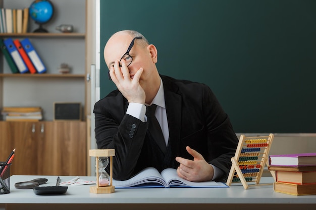 Man teacher wearing glasses sitting at school desk with class register in front of blackboard in classroom explaining lesson looking tired and annoyed