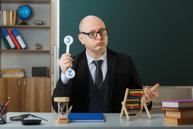 Man teacher wearing glasses sitting at school desk with class register in front of blackboard in classroom holding number plates explaining lesson looking annoyed and displeased