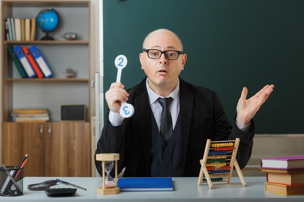 Man teacher wearing glasses sitting at school desk with class register in front of blackboard in classroom holding number plates explaining lesson looking confused having doubts