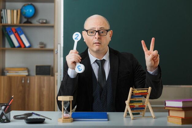 Man teacher wearing glasses sitting at school desk with class register in front of blackboard in classroom holding number plates explaining lesson showing number two