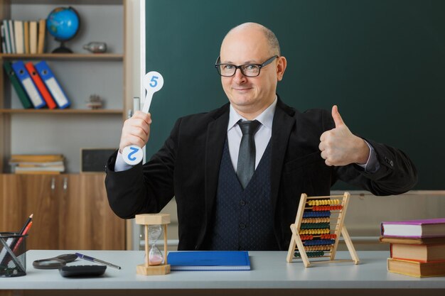 Man teacher wearing glasses sitting at school desk with class register in front of blackboard in classroom holding number plates explaining lesson showing thumb up smiling