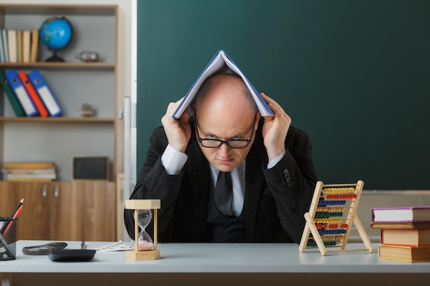 Free photo man teacher wearing glasses sitting at school desk with class register on his head in front of blackboard in classroom explaining lesson frowning