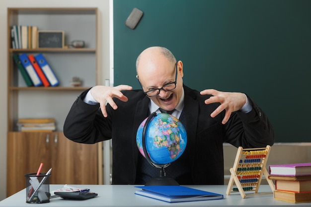 Man teacher wearing glasses sitting with globe at school desk in front of blackboard in classroom explaining lesson angry and frustrated yelling