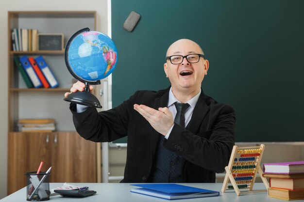 Man teacher wearing glasses sitting with globe at school desk in front of blackboard in classroom explaining lesson happy and excited presenting globe with hand