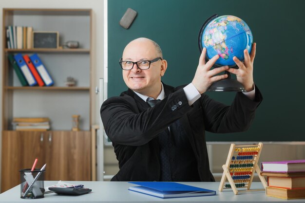 교실에서 칠판 앞 학교 책상에 글로브와 함께 앉아 안경을 쓴 남자 교사는 수업을 설명하고 즐겁게 웃고 기쁘게 생각합니다