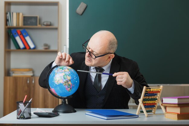 Man teacher wearing glasses sitting with globe at school desk in front of blackboard in classroom explaining lesson holding pointer looking intrigued