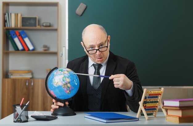 Man teacher wearing glasses sitting with globe at school desk in front of blackboard in classroom explaining lesson holding pointer looking surprised