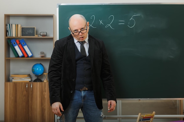 Man teacher wearing glasses standing near blackboard in classroom explaining lesson being tired and overworked