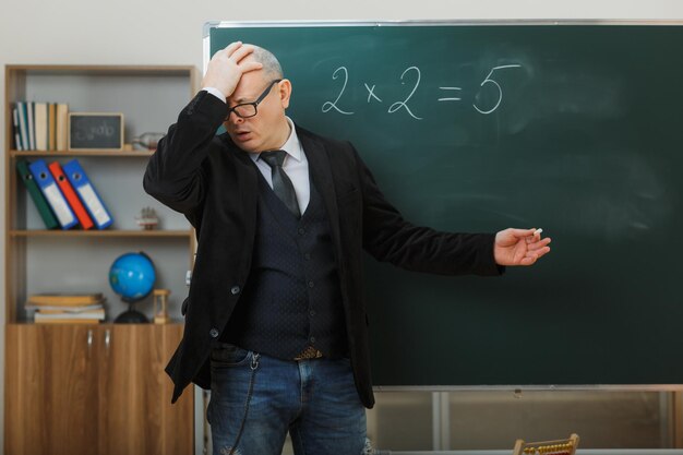 Учитель-мужчина в очках стоит возле доски в классе и объясняет урок, выглядя растерянным, держа руку на голове, забыл что-то важное