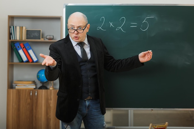 Man teacher wearing glasses standing near blackboard in classroom explaining lesson looking confused raising arms in indignation