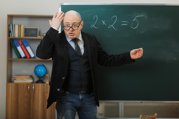 man teacher wearing glasses standing near blackboard in classroom explaining lesson looking disappointed and confused