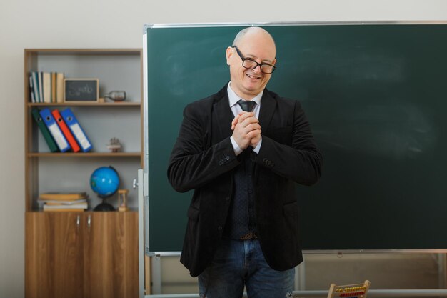 Man teacher wearing glasses standing near blackboard in classroom explaining lesson looking pleased smiling holding hands together