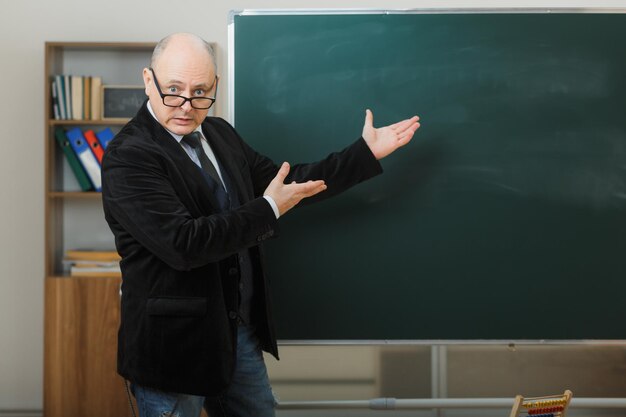 Man teacher wearing glasses standing near blackboard in classroom explaining lesson looking surprised
