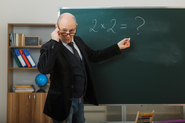 man teacher wearing glasses standing near blackboard in classroom explaining lesson looking surprised