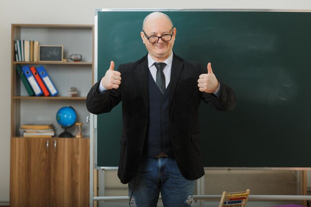 man teacher wearing glasses standing near blackboard in classroom explaining lesson showing thumbs up happy and pleased