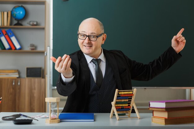 Man teacher wearing glasses with class register sitting at school desk in front of blackboard in classroom happy and positive making come here gesture with hand