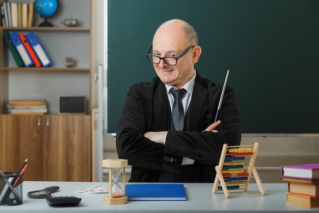 안경을 쓴 남자 교사는 교실에서 칠판 앞 학교 책상에 앉아 수업을 설명하는 포인터를 들고 카메라를 보며 행복하고 기쁘게 웃는다.