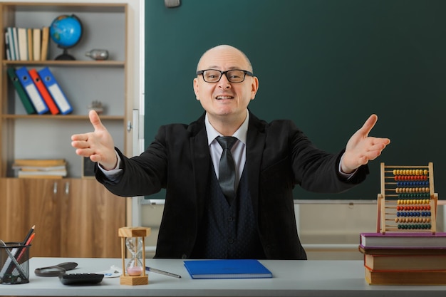 Foto gratuita insegnante uomo che indossa occhiali con registro di classe seduto al banco di scuola davanti alla lavagna in classe guardando la fotocamera felice e compiaciuto ampia apertura mani gesto di benvenuto