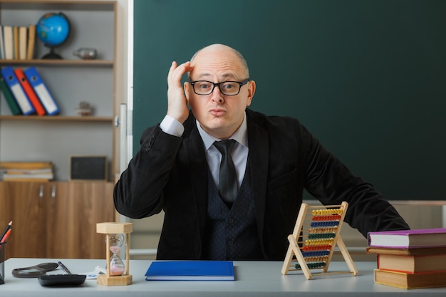 Man teacher wearing glasses with class register sitting at school desk in front of blackboard in classroom looking confused and surprised