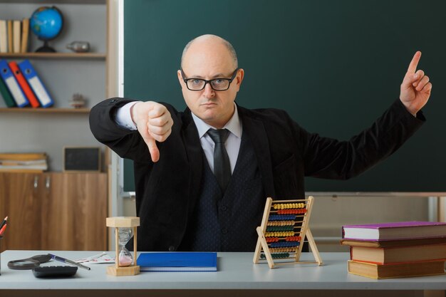 안경을 쓴 남자 교사는 교실에서 칠판 앞 학교 책상에 앉아 있고 검지 손가락으로 가리키는 엄지손가락을 아래로 보여주는 불쾌한 표정을 하고 있다