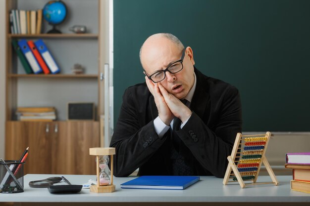 Man teacher wearing glasses with class register sitting at school desk in front of blackboard in classroom looking tired and bored wants to sleep leaning head on palms