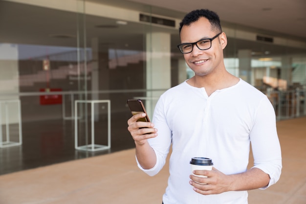 Man texting on phone, holding takeaway coffee, looking at camera