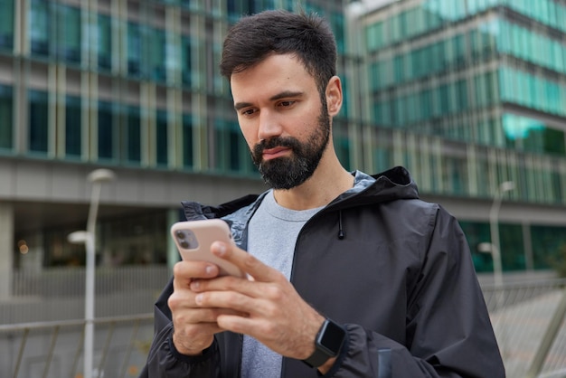 Бесплатное фото Мужчина пользуется мобильным телефоном, делится публикацией о путешествиях после экскурсии по городу, носит черную куртку, устанавливает новое приложение, позирует в городе