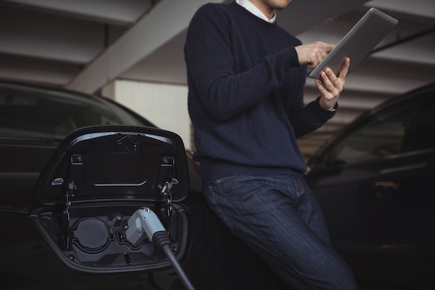 Uomo che utilizza la tavoletta digitale durante la ricarica dell'auto elettrica