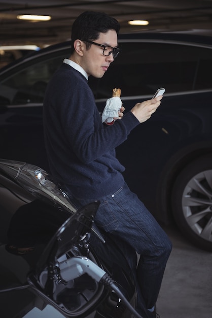 Free photo man using mobile phone while charging electric car