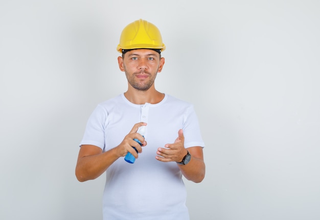 Man using spray bottle for cleaning hands in t-shirt, helmet and looking careful, front view.
