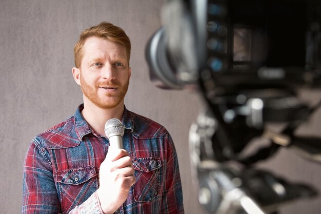 Man videoing himself and holding microphone in his hand. Stunning young man with red hair wearing shirt standing in front of video camera speaking into microphone.