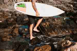 Free photo man walking on stone beach with surf board near water