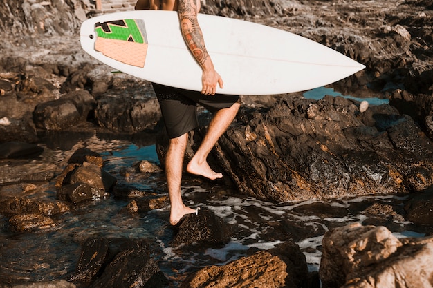 Man walking on stone beach with surf board near water